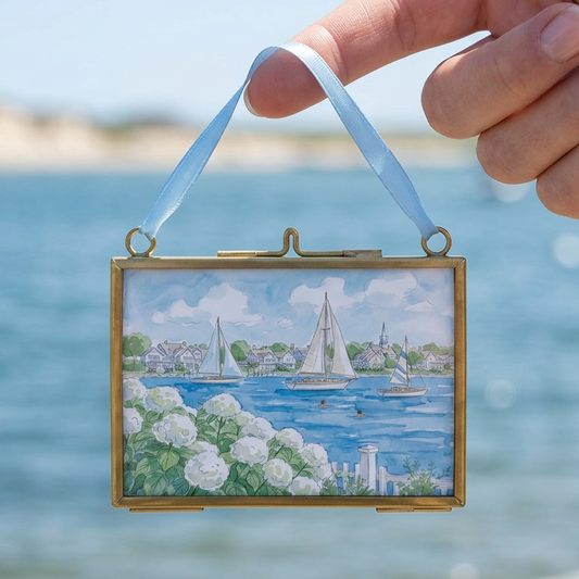 Hand holding a small framed artwork of a coastal scene with boats against a blurred ocean background.